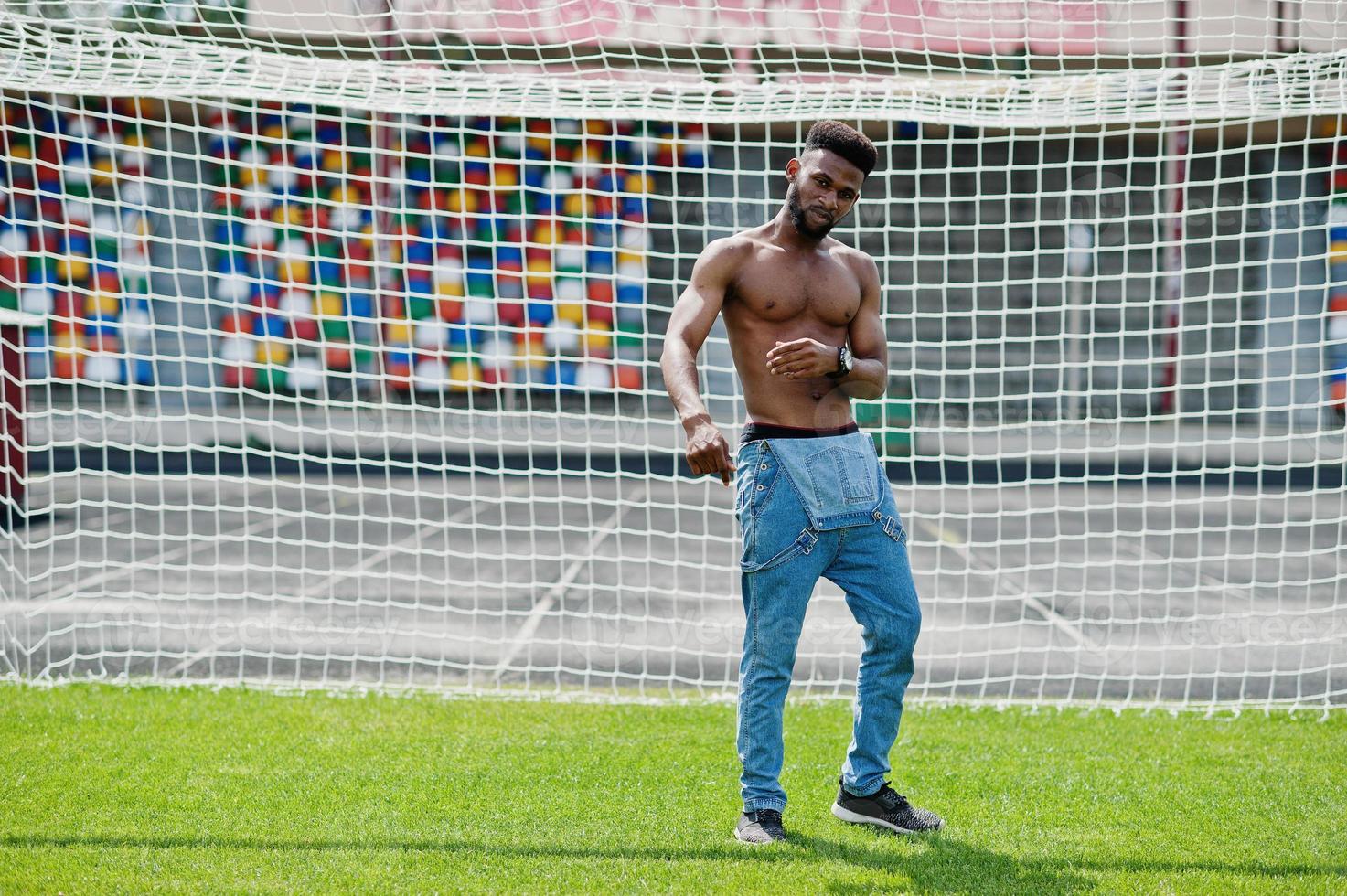 Handsome sexy african american muscular bare torso man at jeans overalls posed at green grass agasinst football gates of stadium field. Fashionable black man portrait. photo