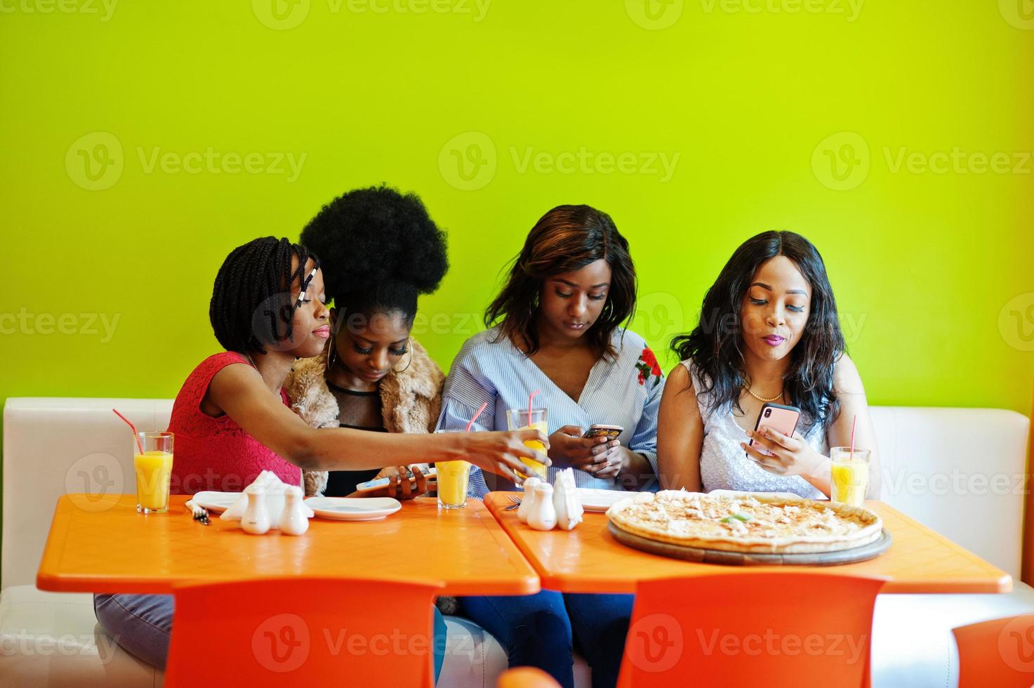 cuatro jóvenes africanas en un restaurante de comida rápida de colores brillantes haciendo fotos de pizza en sus teléfonos móviles.