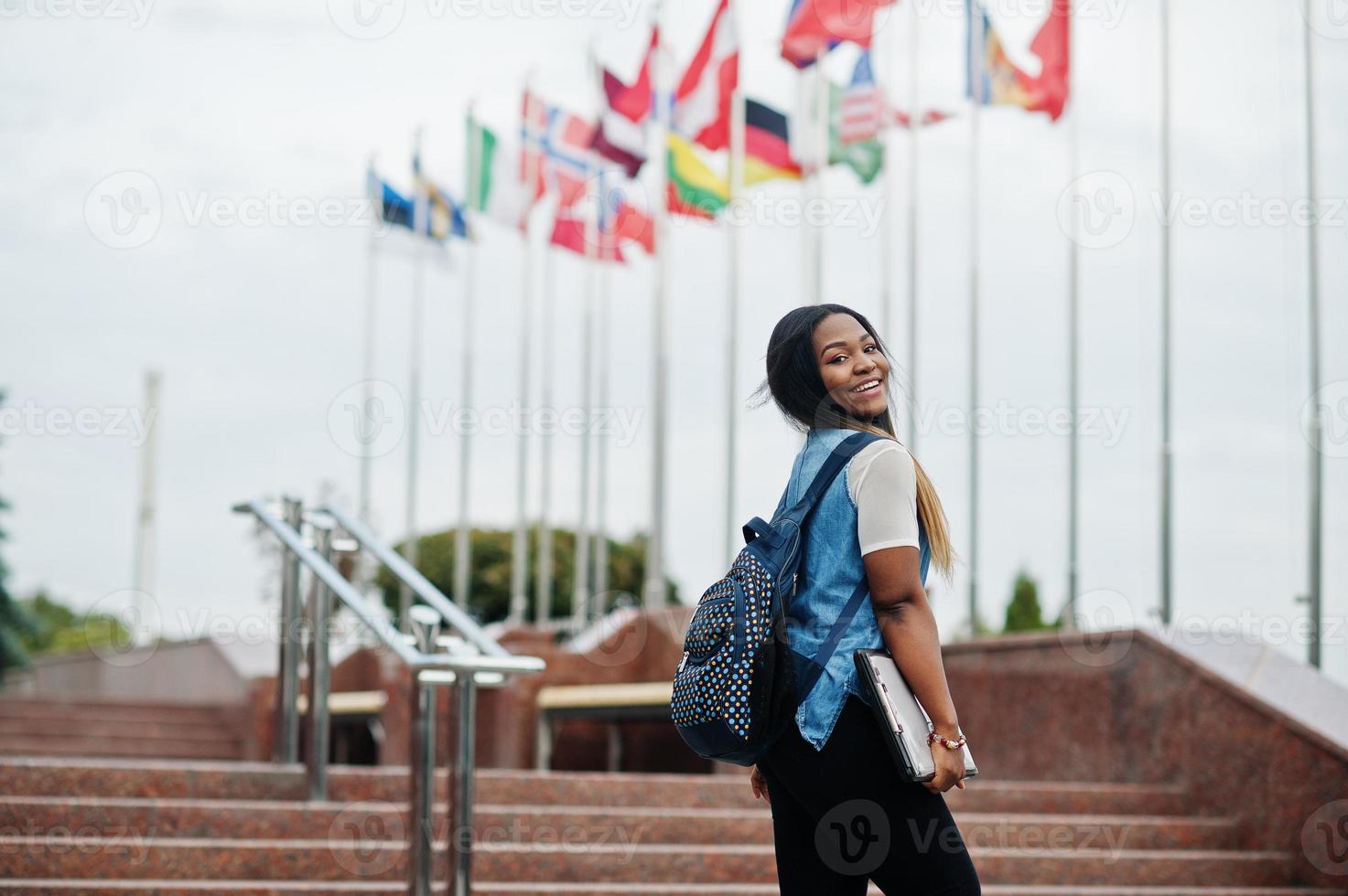 African student female posed with backpack and school items on yard of university, against flags of different countries. photo