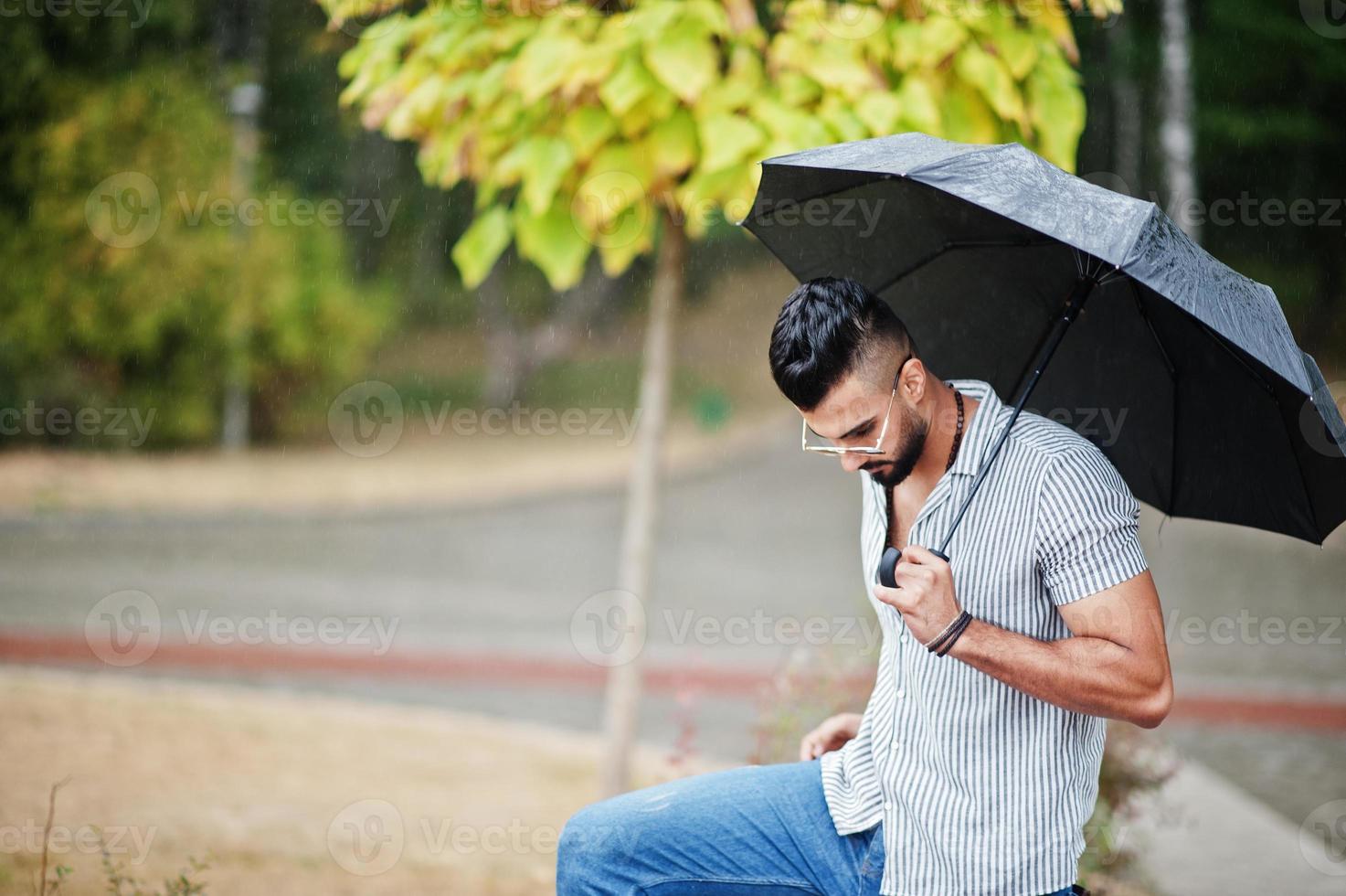 Fashionable tall arab beard man wear on shirt, jeans and sunglasses with umbrella posed at rain on park square. photo