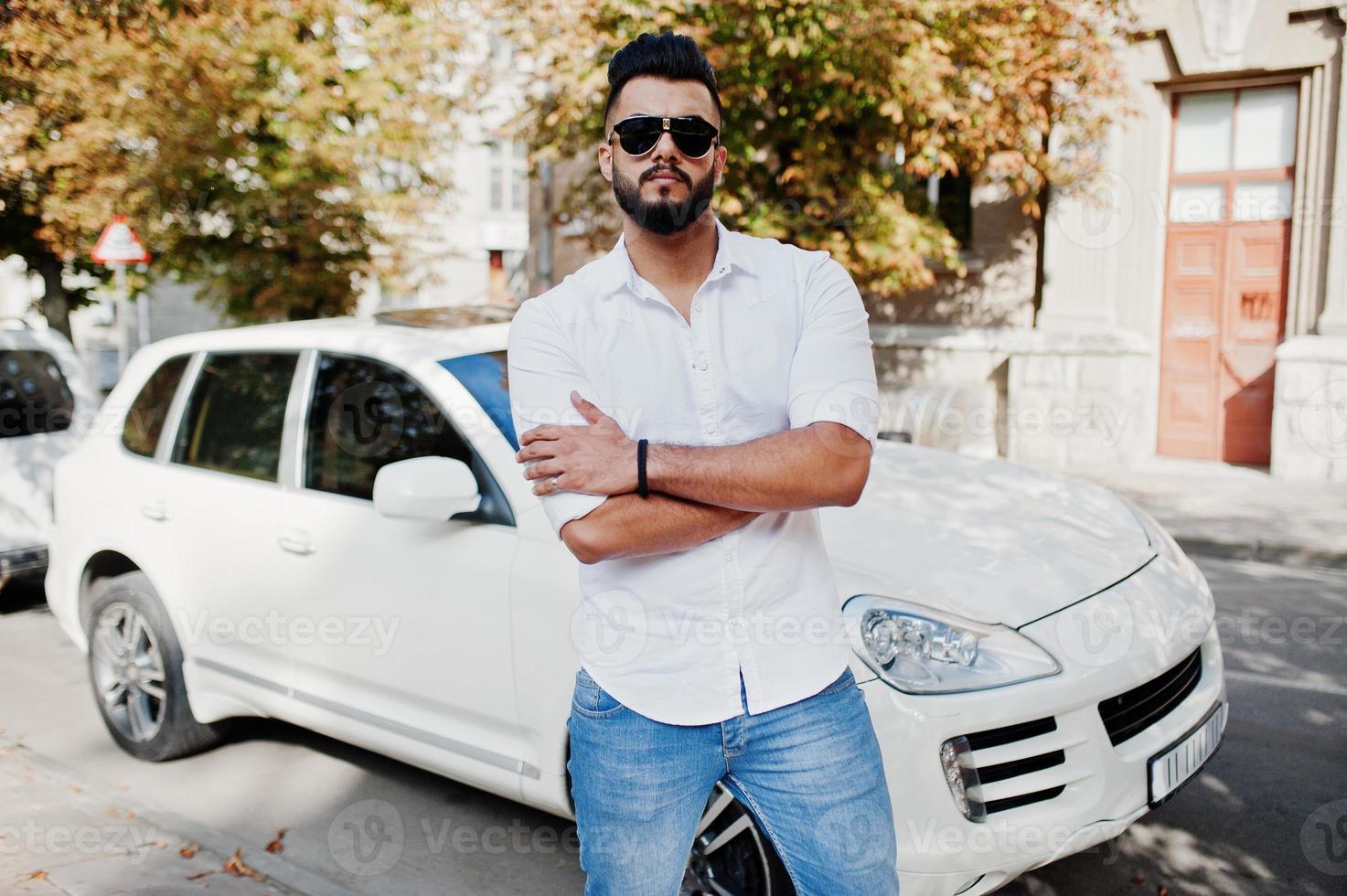 Elegante modelo de hombre árabe alto con camisa blanca, jeans y gafas de sol en la calle de la ciudad. chico árabe rico y atractivo con barba contra un auto todoterreno blanco. foto