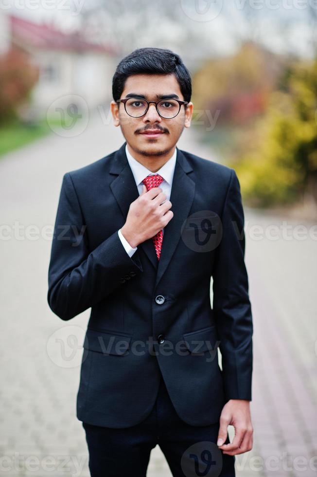 Indian young man at glasses, wear on black suit with red tie posed outdoor. photo