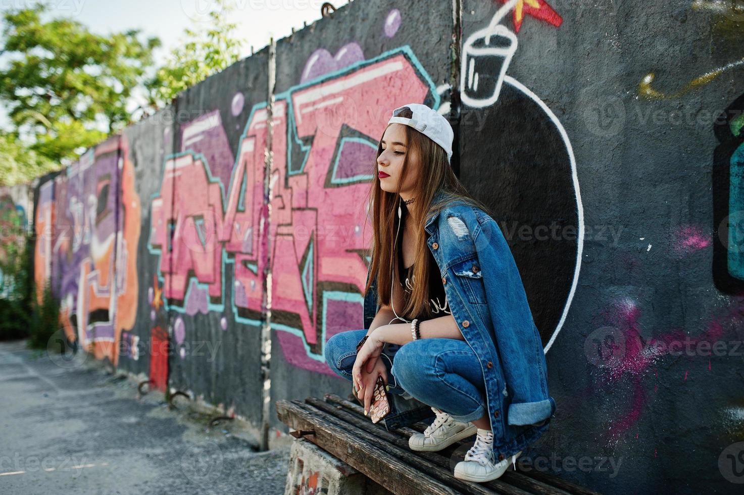 Stylish casual hipster girl in cap and jeans wear listening music from headphones of mobile phone against large graffiti wall with bomb. photo