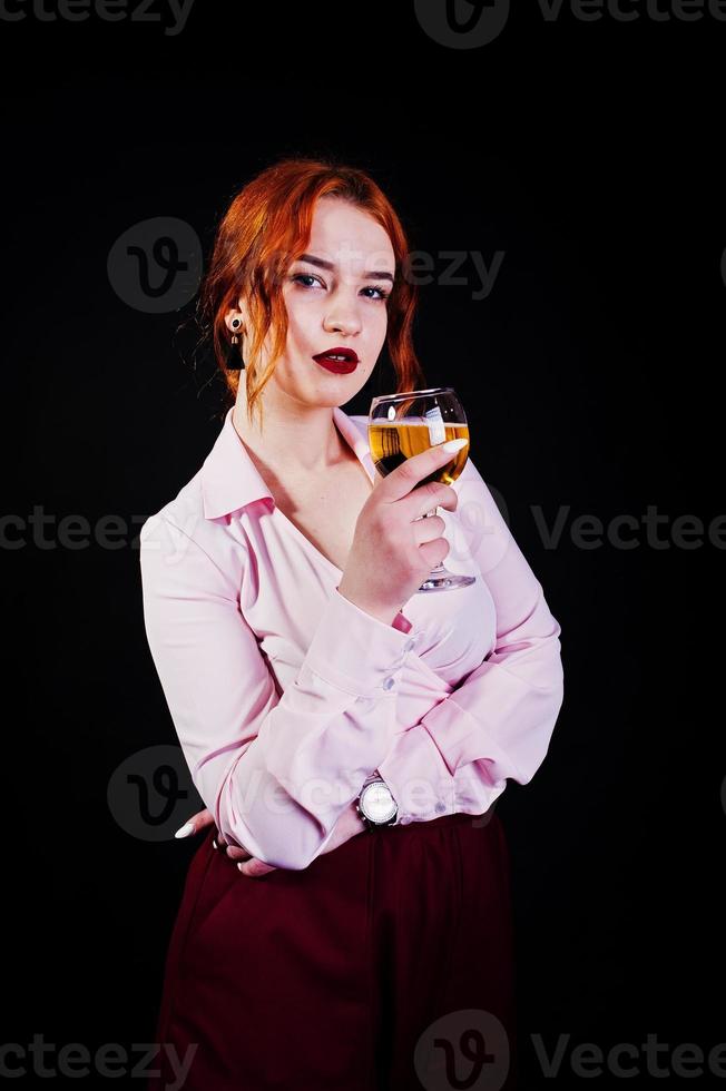 Gorgeous red haired girl in pink blouse and red skirt with glass of wine at hand isolated on black. photo