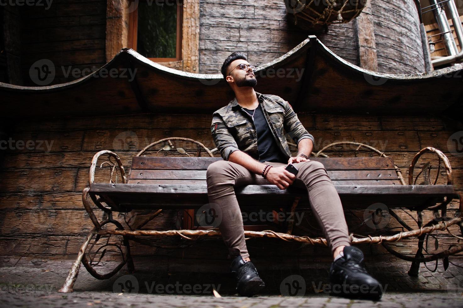 Awesome beautiful tall ararbian beard macho man in glasses and military jacket posed outdoor, sitting on bench against wooden house. photo