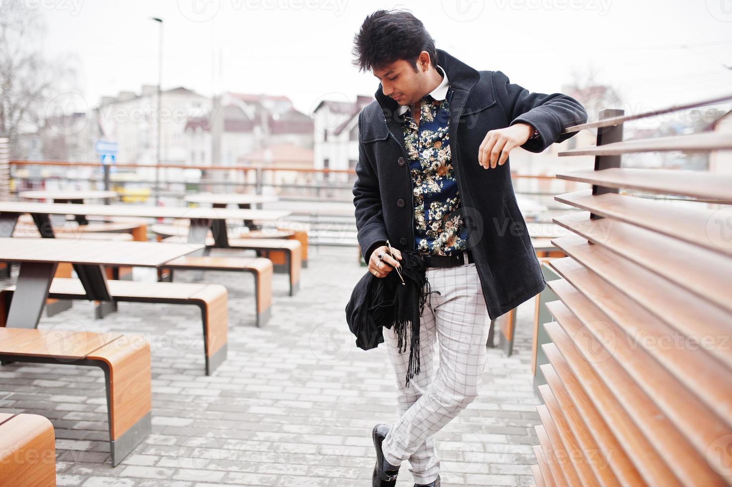 atractivo y apuesto hombre indio con estilo vestido con un abrigo negro parado en la terraza del moderno café salón en su descanso de trabajo. foto