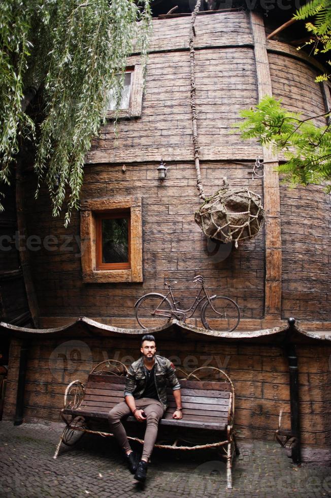 Awesome beautiful tall ararbian beard macho man in glasses and military jacket posed outdoor, sitting on bench against wooden house. photo