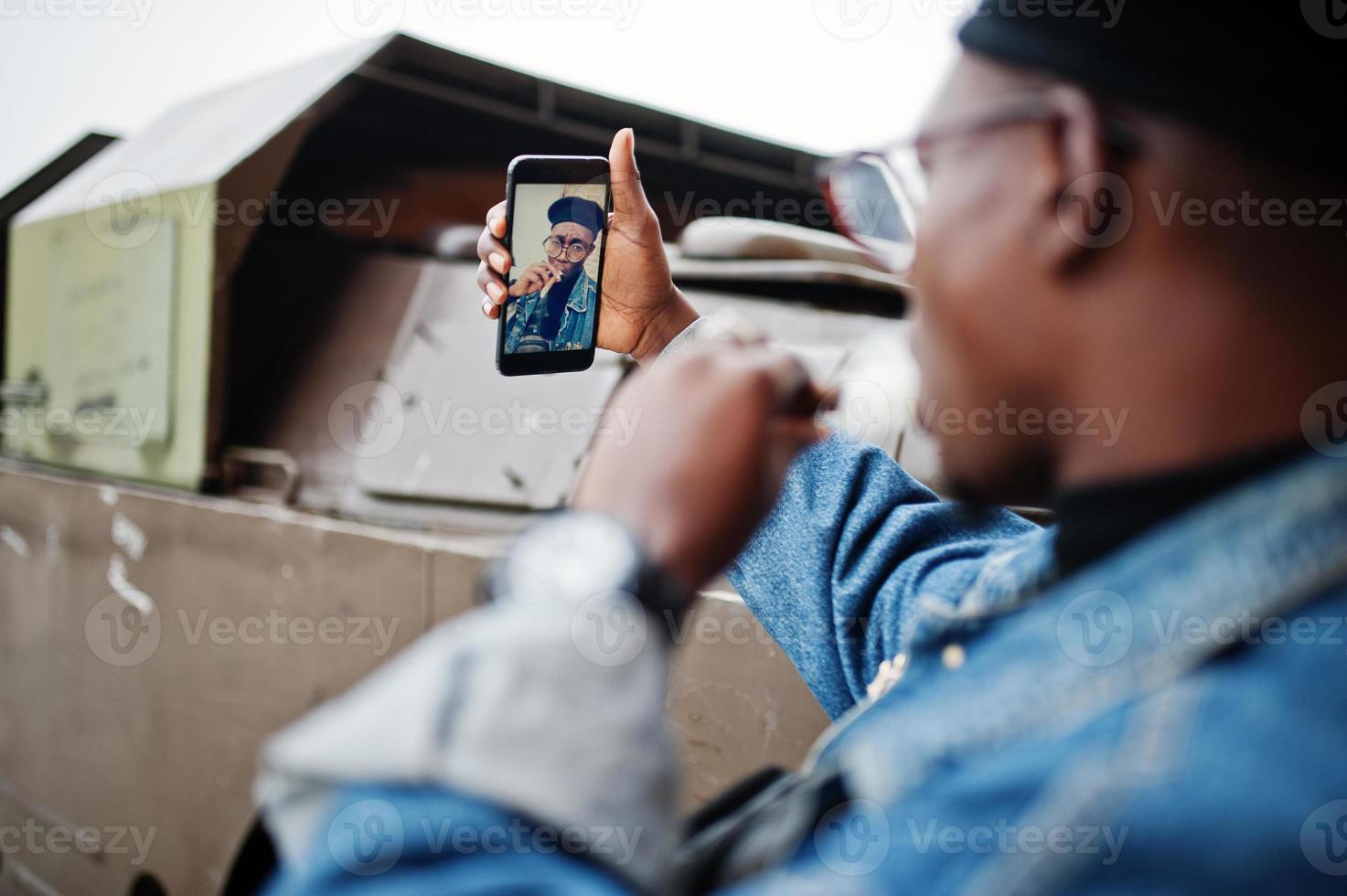 African american man in jeans jacket, beret and eyeglasses, smoking cigar and posed against btr military armored vehicle, making selfie on phone. Focus on screen of cellphone. photo