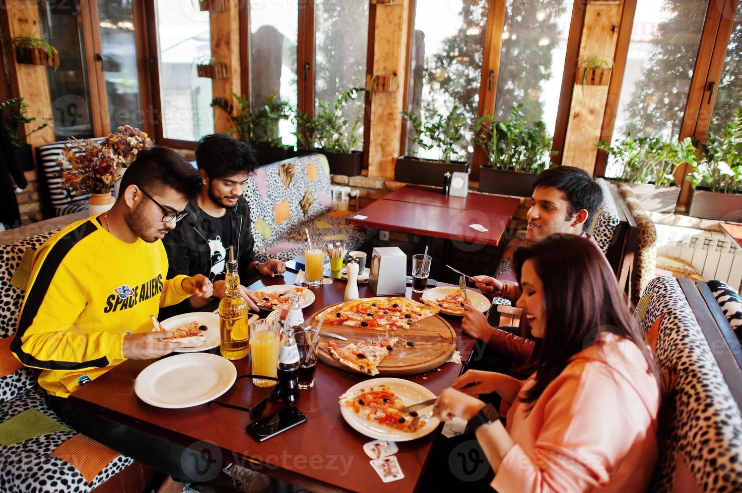 Group of asian friends eating pizza during party at pizzeria. Happy indian people having fun together, eating italian food and sitting on couch. photo