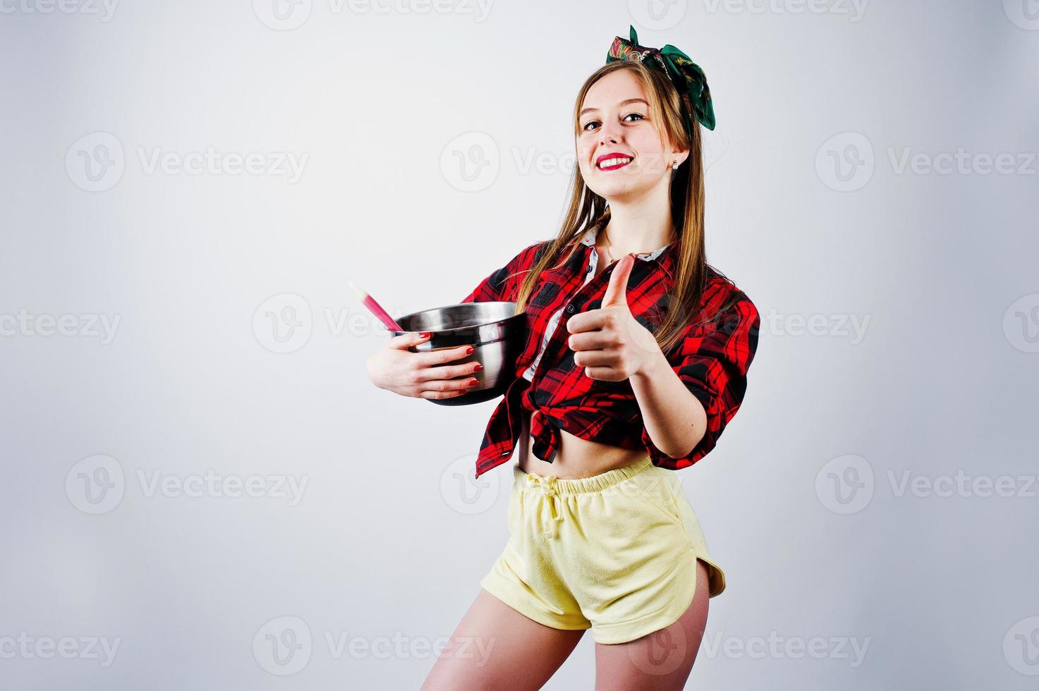 Young funny housewife in checkered shirt and yellow shorts pin up style with saucepan and kitchen spoon isolated on white background. photo