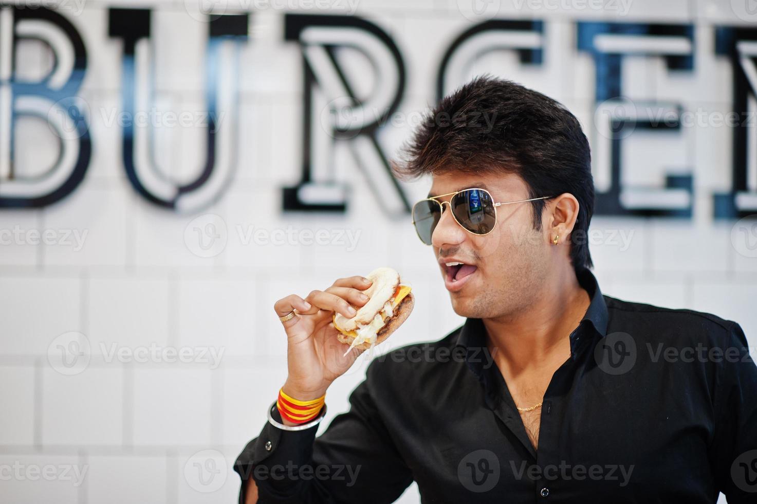 un hombre indio con estilo con gafas de sol en un café de comida rápida come una hamburguesa contra un cartel de hamburguesa en la pared. foto