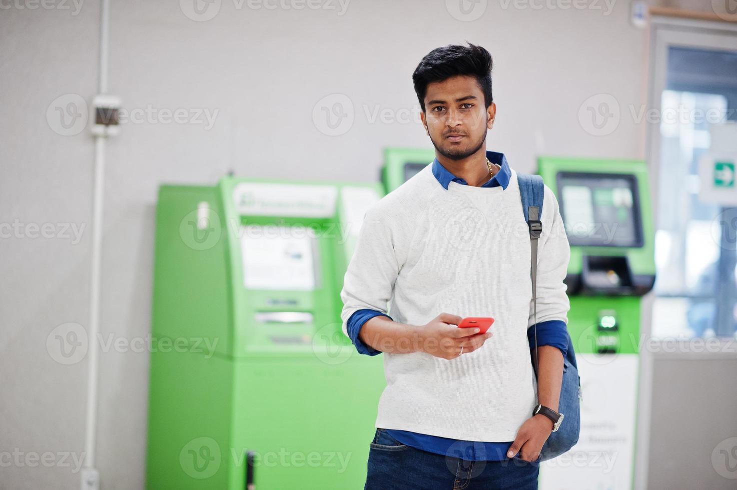 Young stylish asian man with mobile phone and backpack against row of green ATM. photo