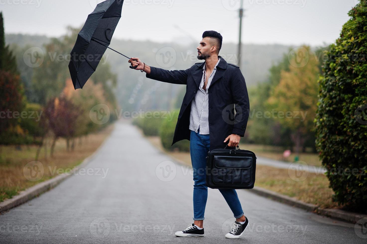 Fashionable tall arab beard man wear on black coat with umbrella and bag case posed at rain weather day. photo