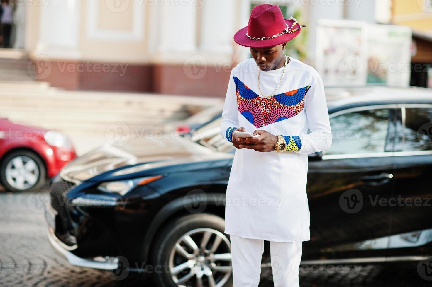 Stylish trendy afro france man in red hat and white outfit posed at autumn day. Black african model business guy with mobile phone at hands against luxury car. photo