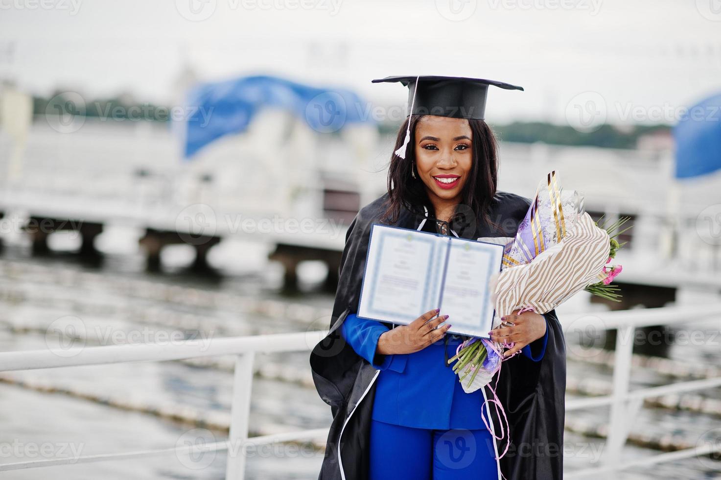 chica estudiante afroamericana en vestido de graduación negro con diploma, en traje azul posado. foto