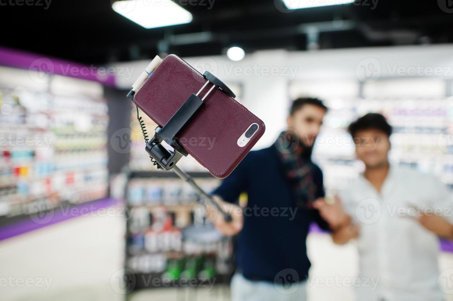 Close up photo of mobile phone on monopod stick against two indians mans customer buyer aking selfie. South asian peoples and technologies concept. Cellphone shop.