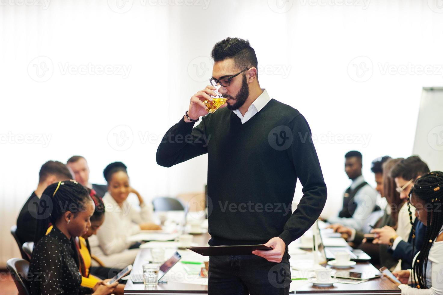 Face of handsome arabian business man, holding clipboard and glasses of beverage on the background of business peoples multiracial team meeting, sitting in office table. photo