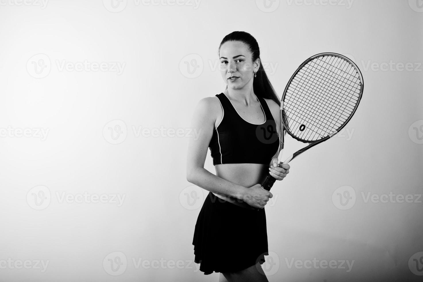 Black and white portrait of beautiful young woman player in sports clothes holding tennis racket while standing against white background. photo