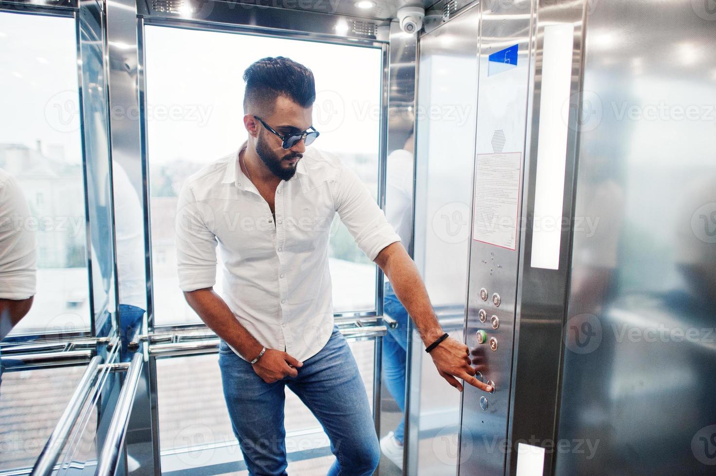 elegante modelo de hombre árabe alto con camisa blanca, jeans y gafas de sol posadas en el interior del ascensor. foto