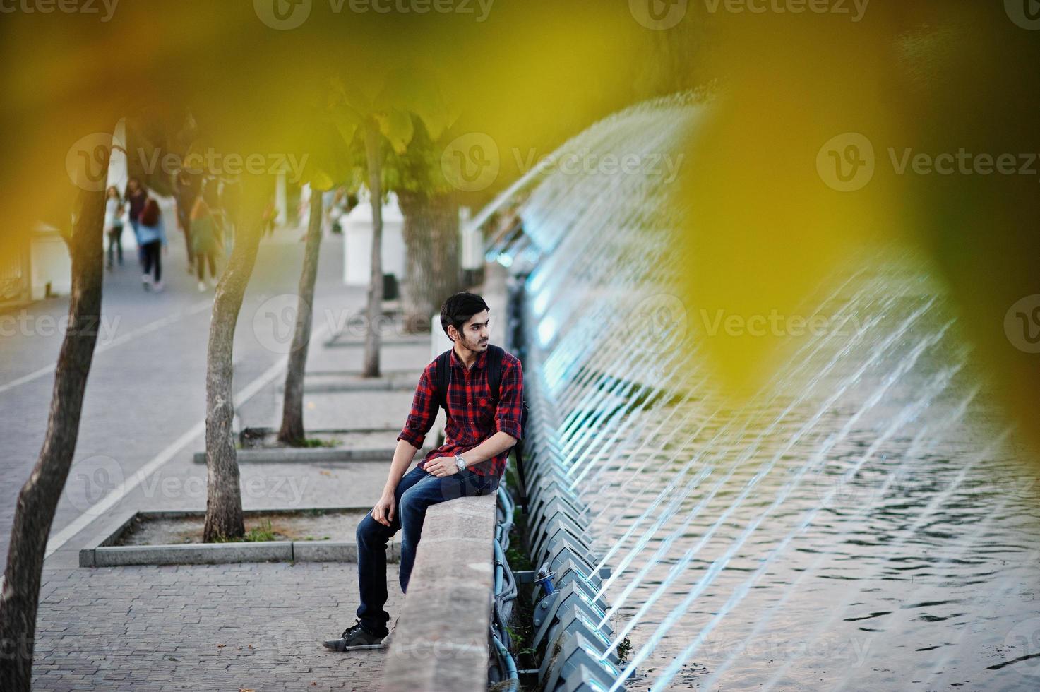 joven estudiante indio con camisa a cuadros y jeans con mochila posada en la ciudad nocturna contra las fuentes. foto