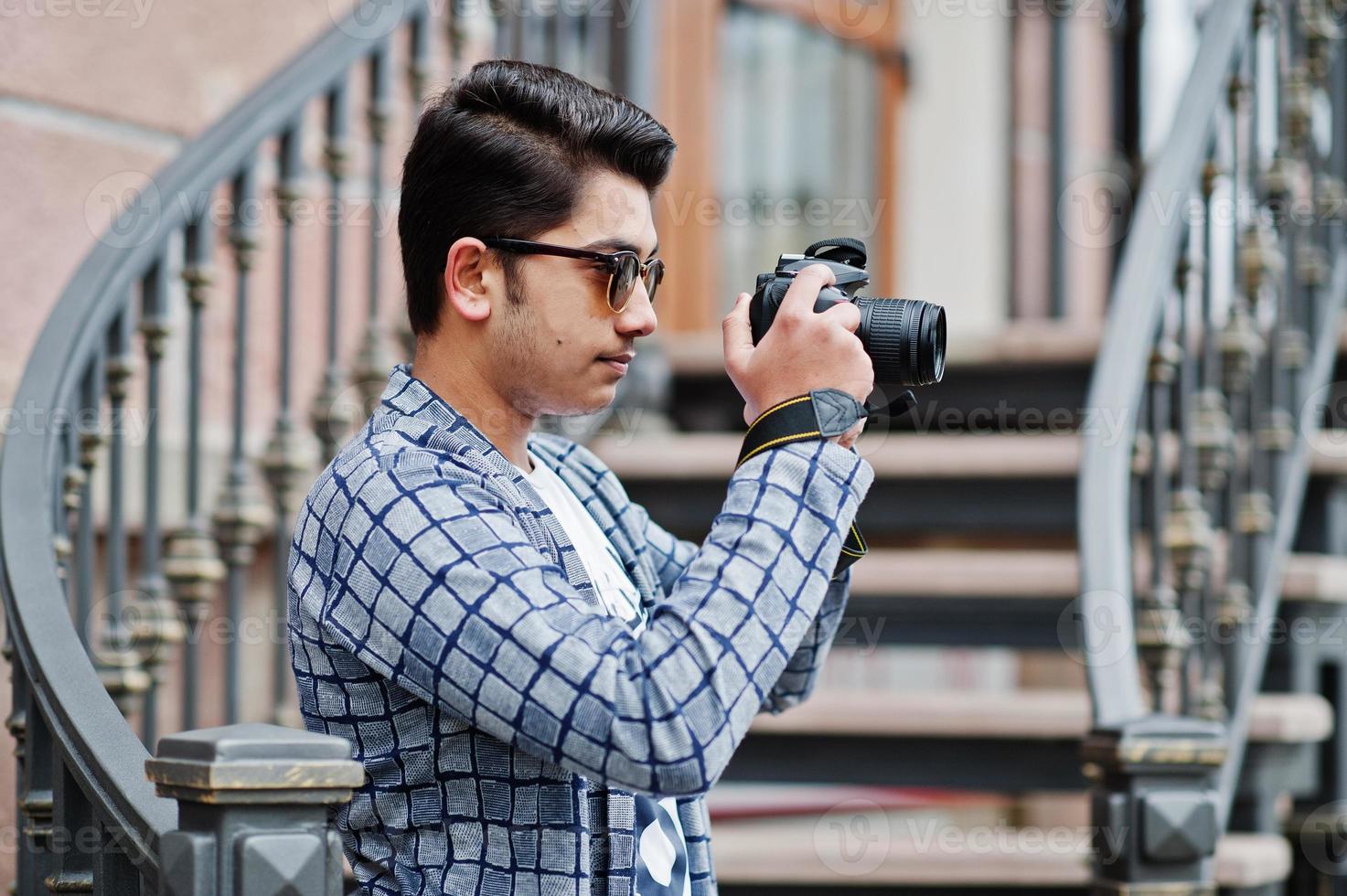 Stylish indian young man photographer at sunglasses wear casual posed outdoor against iron stairs with dslr photo camera at hands.