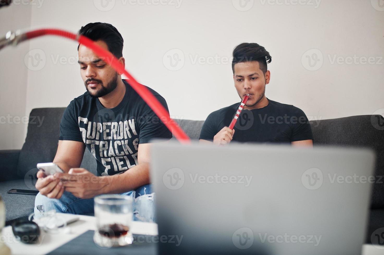 Two handsome and fashionable indian mans relaxing at room and smoking hookah. One of them looking at mobile phone. photo