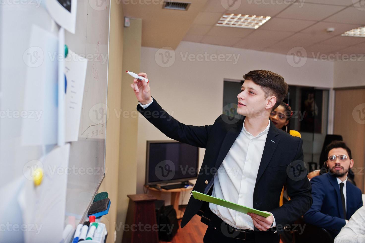 Mixed race business coach presenting report standing near whiteboard pointing on sales statistic shown on diagram and chart teach diverse company members gathered together in conference room. photo