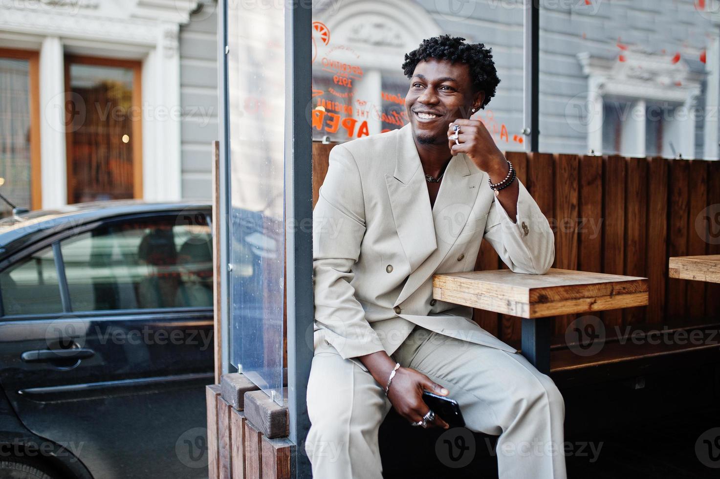 Stylish afro man in beige old school suit sit at table on street. Fashionable young African male in casual jacket on bare torso. photo