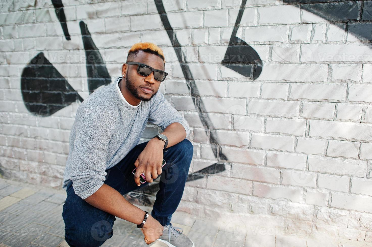 Stylish african american boy on gray sweater and black sunglasses posed at street. Fashionable black guy against graffity wall. photo