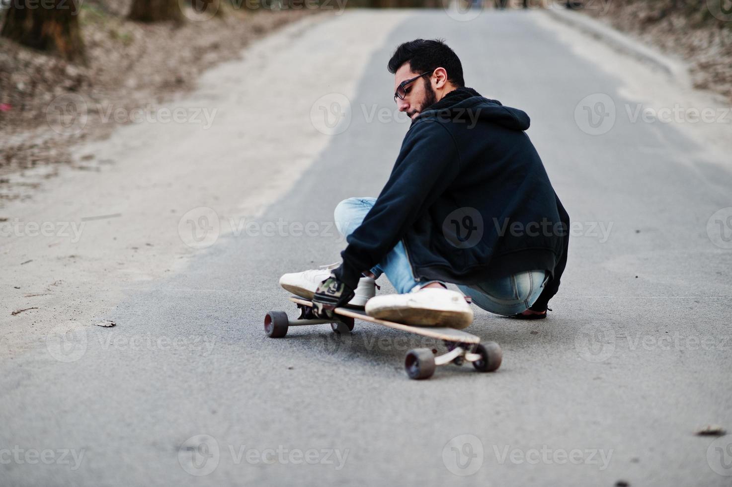Street style arab man in eyeglasses with longboard longboarding down the road. photo