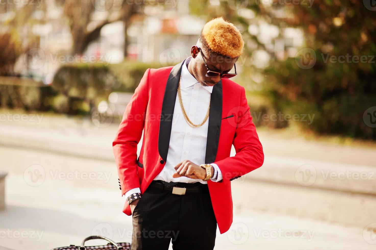 Fashion african american man model at red suit, with highlights hair and handbag posed at street and looking at his watches. photo