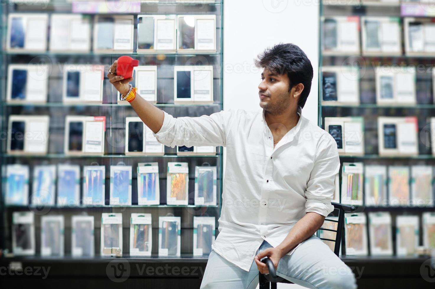 Indian man customer buyer at mobile phone store with wireless earphones and speaker sitting on chair. South asian peoples and technologies concept. Cellphone shop. photo