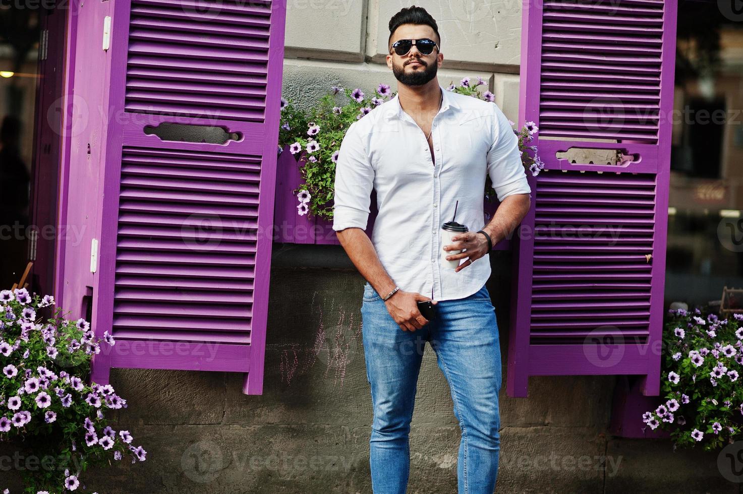 Stylish tall arabian man model in white shirt, jeans and sunglasses posed at street of city. Beard attractive arab guy with cup of coffee against purple windows. photo