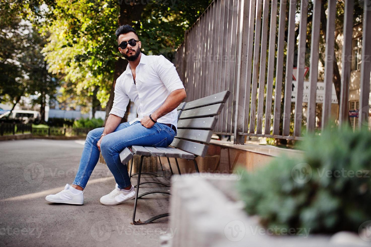 Stylish tall arabian man model in white shirt, jeans and sunglasses posed at street of city. Beard attractive arab guy sitting on bench. photo