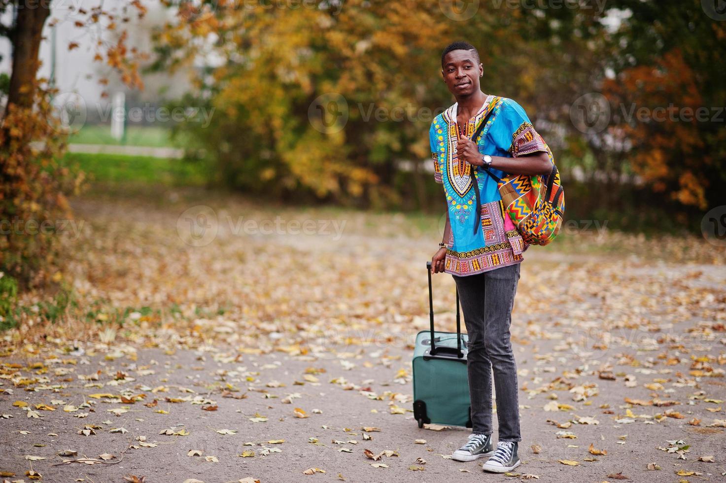 African man in africa traditional shirt on autumn park with backpack and suitcase. Emigrant traveler. photo