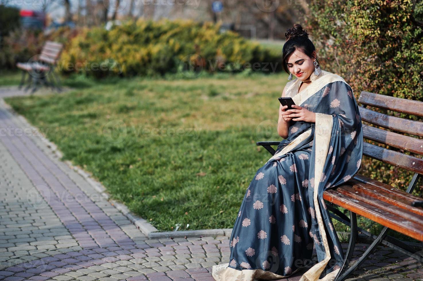 Elegant brunette south asian indian girl in saree sitting on bench with mobile phone at hands. photo