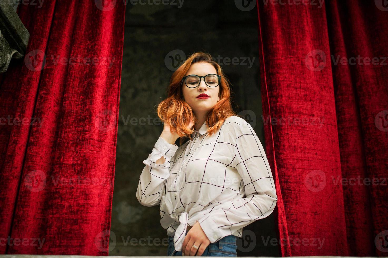 Attractive redhaired woman in eyeglasses, wear on white blouse posing at arch of open red curtains. photo