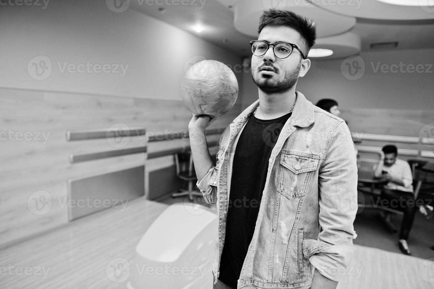 Stylish asian man in jeans jacket and glasses standing at bowling alley with ball at hand. photo