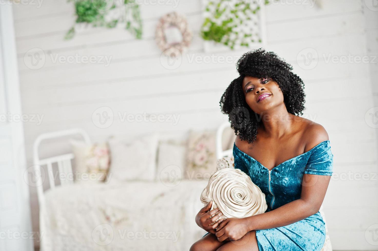 Afican american women in brilliant glitter sequins turqoise dress sitting on chair at white room with decorative rose at hands. photo