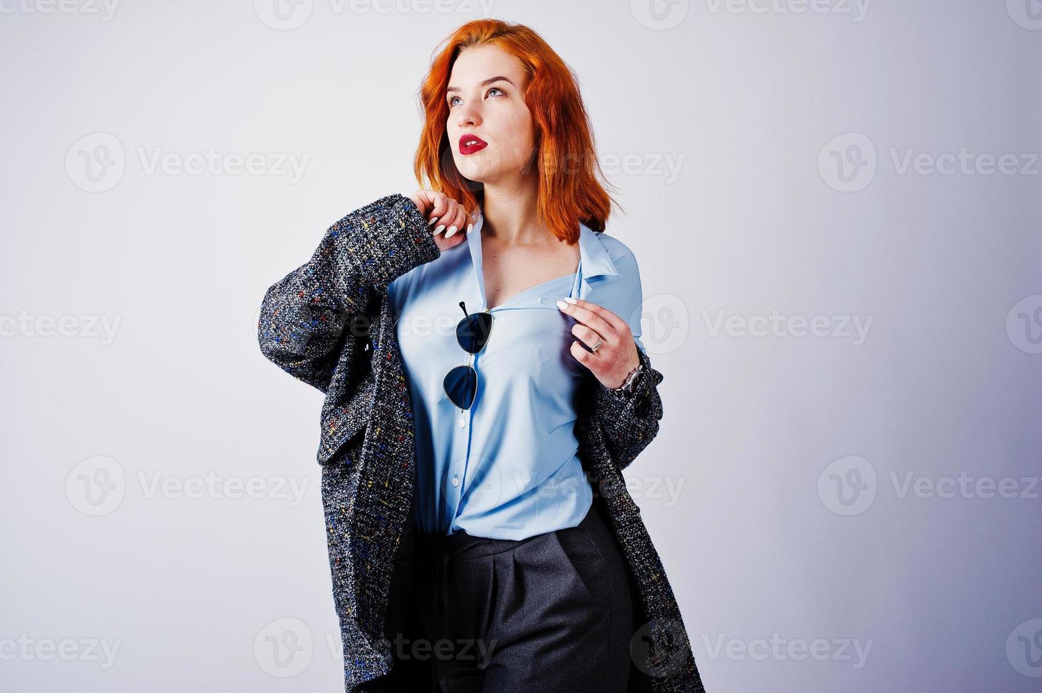 Portrait of a fantastic redheaded girl in blue shirt, grey overcoat posing with sunglasses in the studio. photo