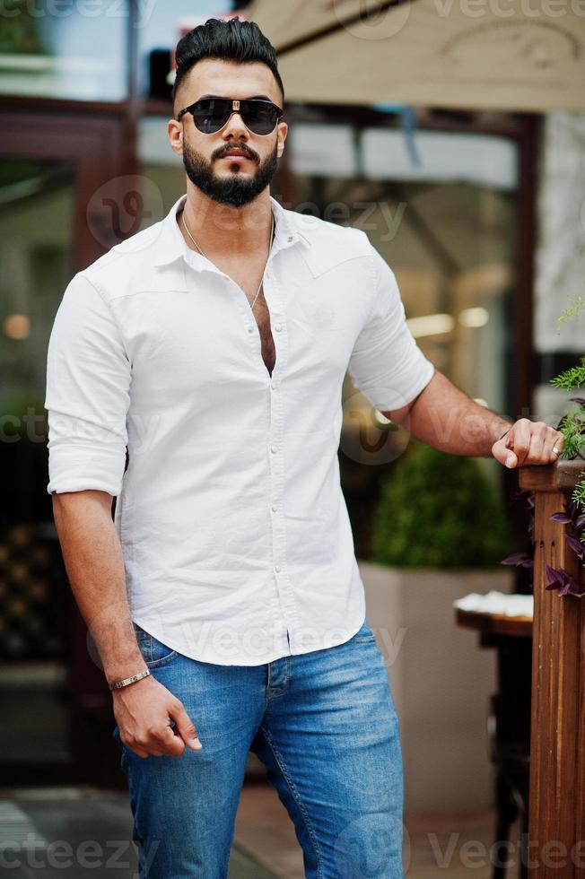 Elegante modelo de hombre árabe alto con camisa blanca, jeans y gafas de sol en la calle de la ciudad. barba chico árabe atractivo. Foto de stock en Vecteezy