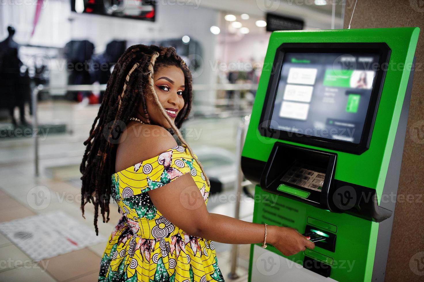 linda chica afroamericana de pequeña altura con rastas, vestida con un vestido amarillo de color, contra un cajero automático con tarjeta de crédito a mano. foto