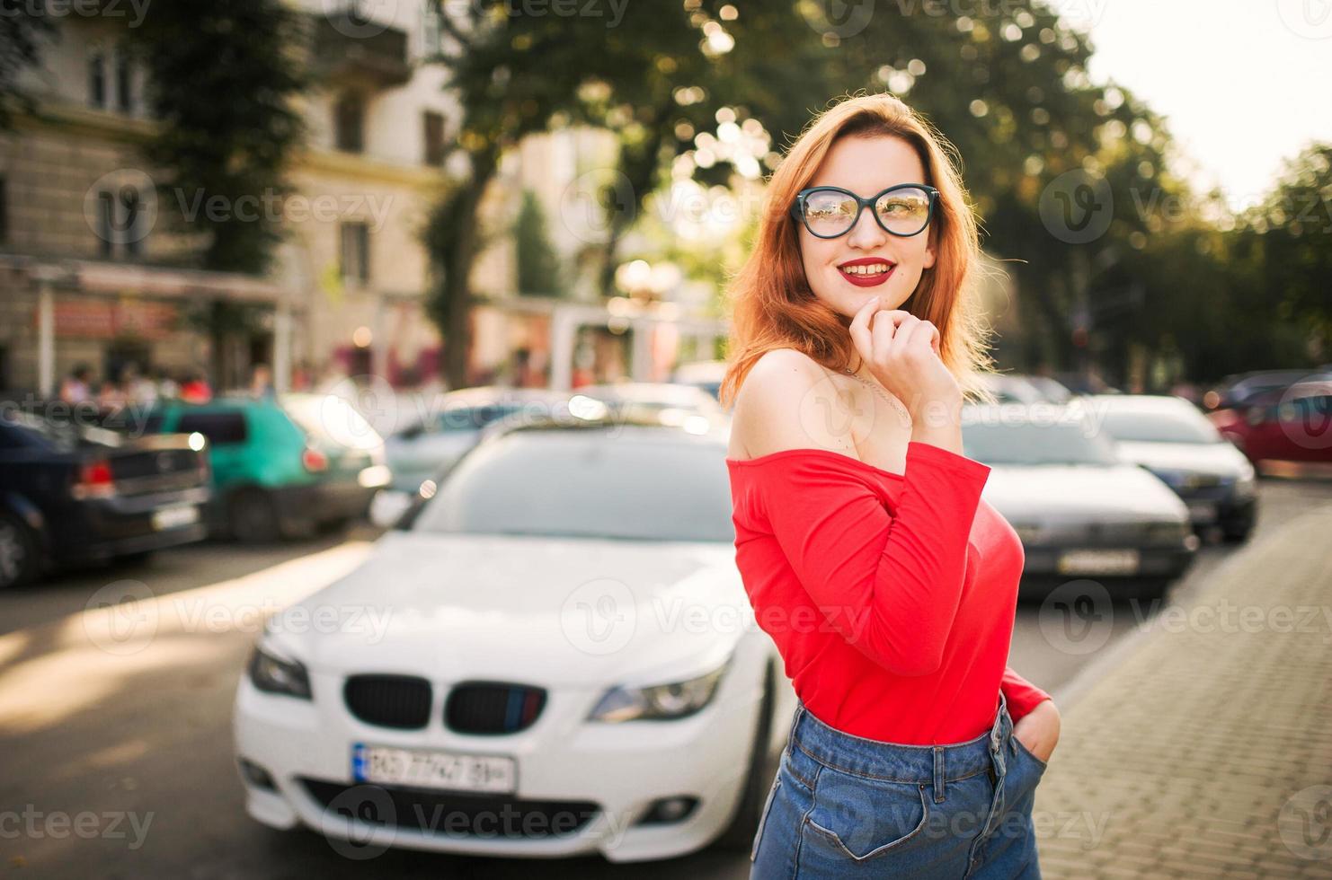 atractiva mujer pelirroja con anteojos, blusa roja y falda de jeans posando en la calle contra un auto deportivo blanco. foto