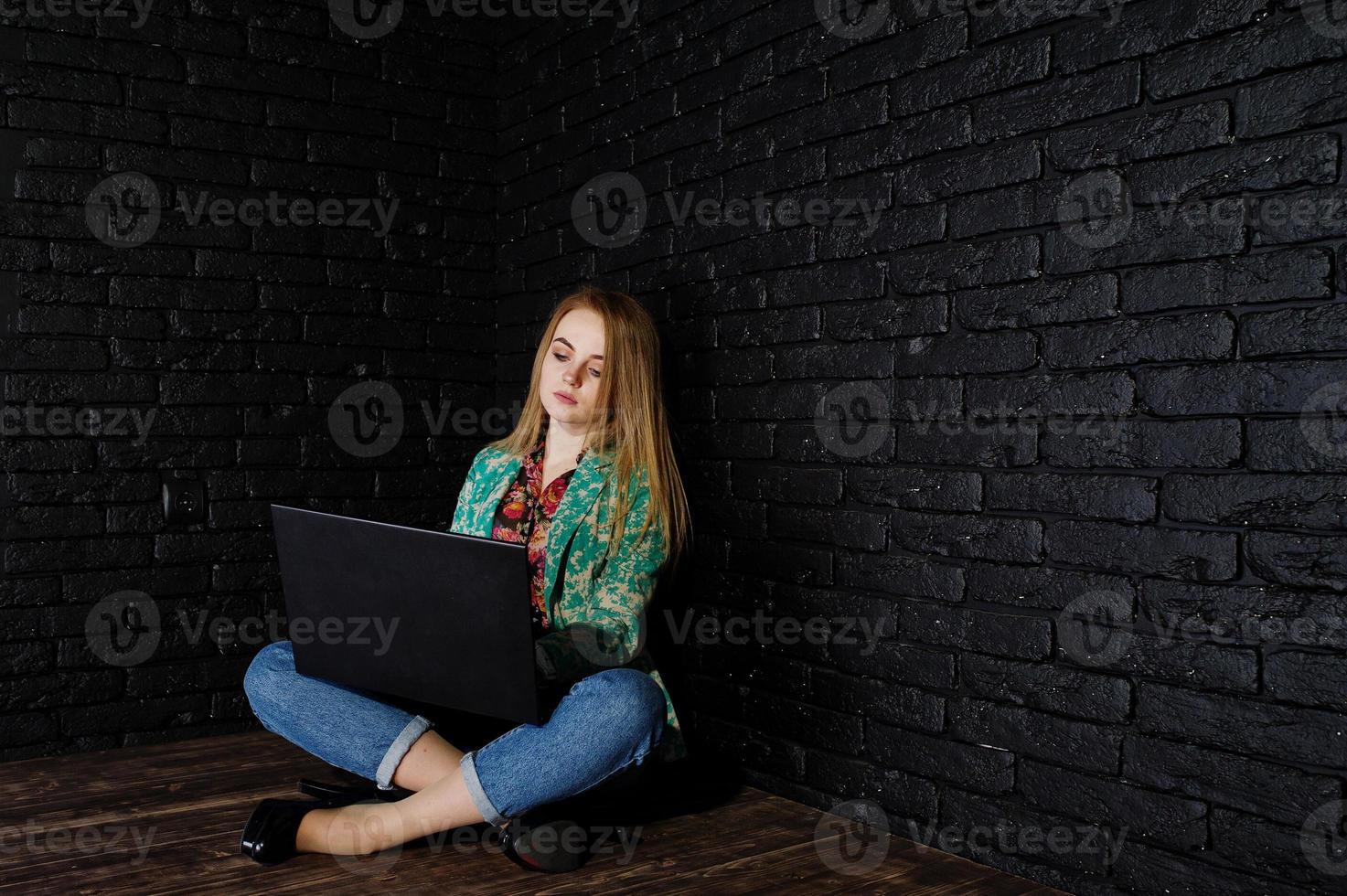 Stylish blonde girl in jacket and jeans with laptop against brick black wall at studio. photo