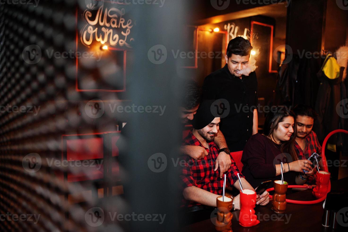 grupo de amigos indios divirtiéndose y descansando en el club nocturno, bebiendo cócteles y fumando narguile, mirando teléfonos móviles. foto