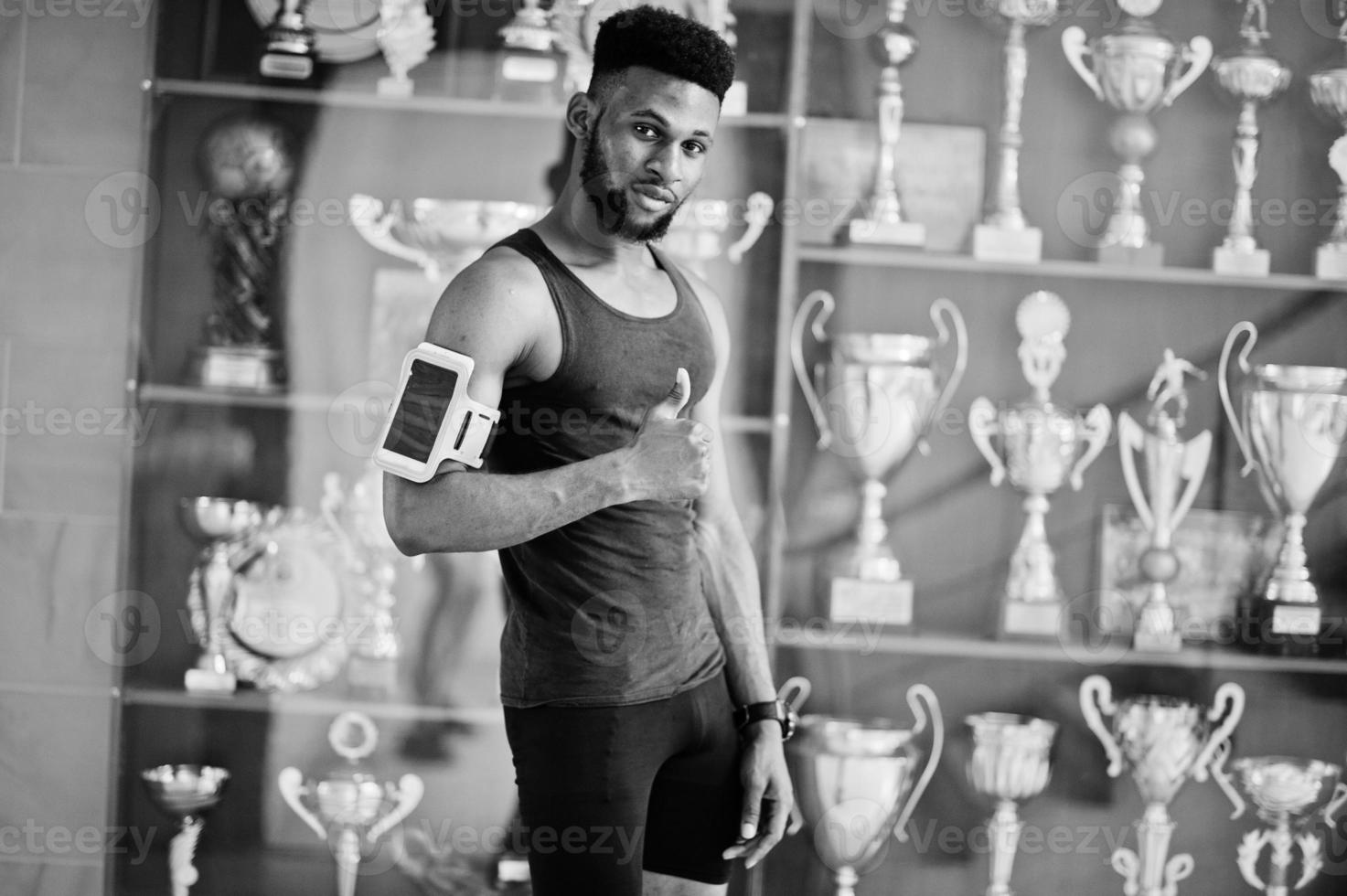African american male athlete sexy sport man with running sports arm case for mobile phone, posed against many golden cups. photo