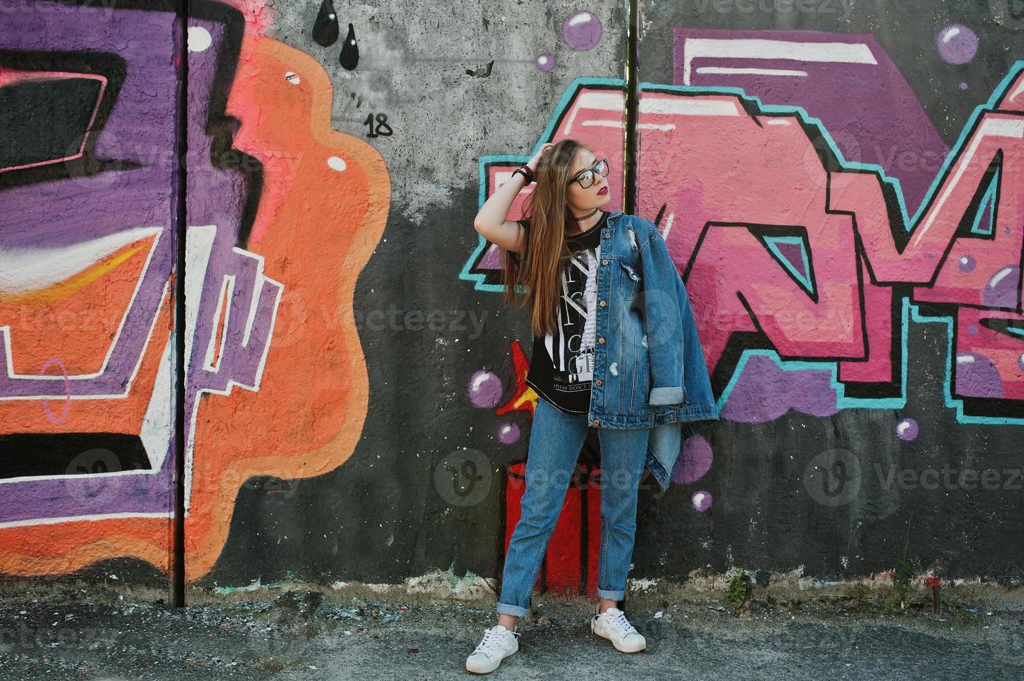 Stylish casual hipster girl in jeans wear and glasses against large graffiti wall. photo