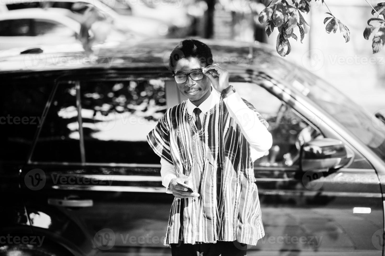 African businessman in traditional clothes and glasses with mobile phone against black car suv. Rich africans people. photo