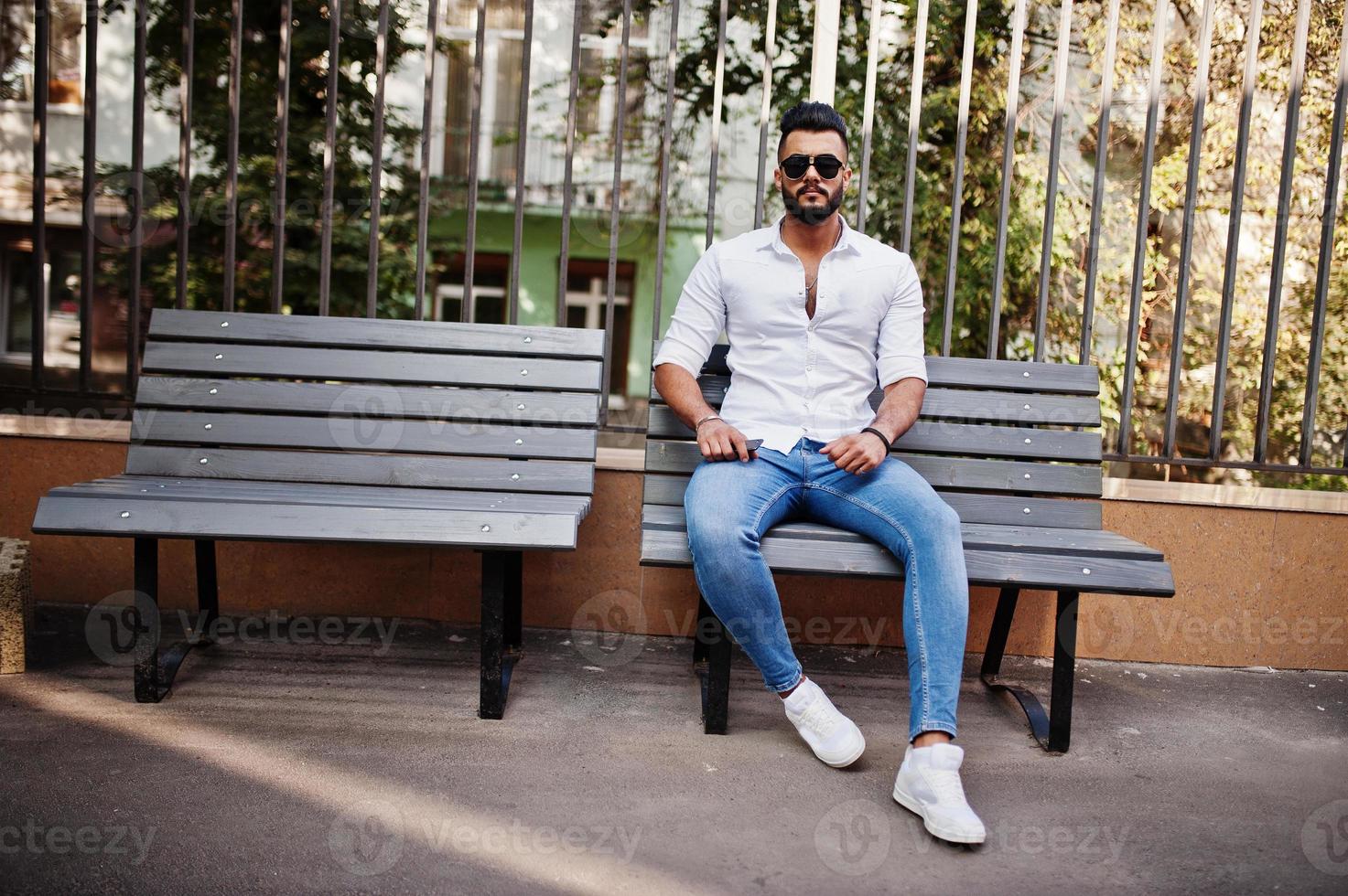 Stylish tall arabian man model in white shirt, jeans and sunglasses posed at street of city. Beard attractive arab guy sitting on bench. photo