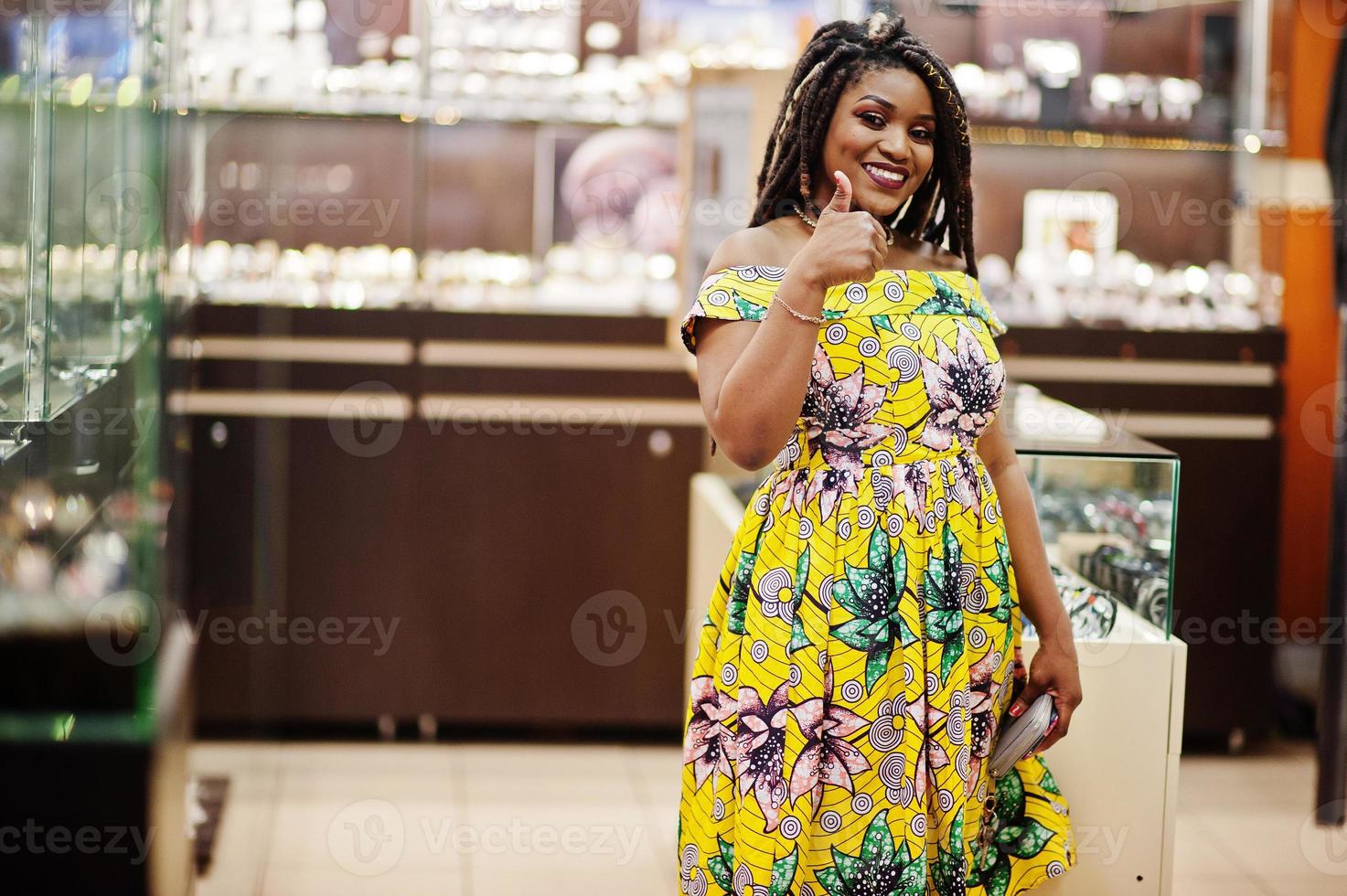 linda chica afroamericana de pequeña altura con rastas, vestida con un vestido amarillo de color, en una tienda de relojes en el centro comercial. foto